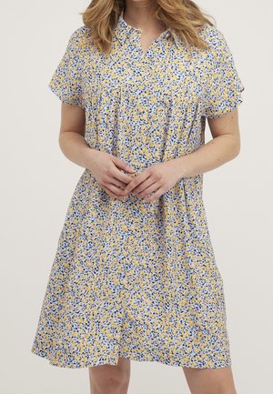 Kvinde iført kortærmet, knælængde kjole med gul, blå og hvidt blomsterprint, hænder løst foldet foran.