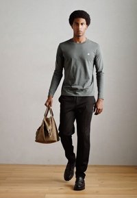 Maglietta grigia a maniche lunghe, pantaloni neri e scarpe nere; tiene in mano una borsa marrone. Tessuto liscio con una vestibilità aderente, in piedi su un pavimento di legno.