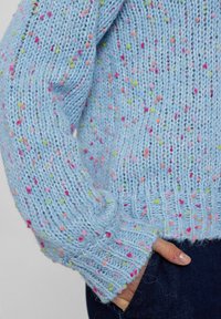 Jasnoniebieski sweter dzianinowy z kolorowymi flekami. Żebrowane mankiety i dół. Tekstura jest puszysta, o luźnym kroju i widocznych detalach ściegów.