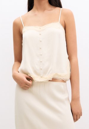 Femme portant un caraco en satin couleur crème avec des boutons à l'avant, une bordure en dentelle, et une jupe assortie sur un fond blanc uni.