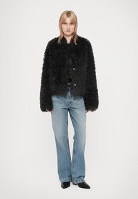 Person with blonde hair wearing a black fuzzy jacket, light blue wide-leg jeans, and black pointed shoes standing against a plain background.