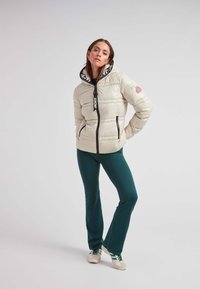 Giacca a vento in un tenue beige chiaro, con chiusura lampo nera e cappuccio. Accentata da una patch con logo rosa e fodera interna a motivo. Abbinata a pantaloni verdi.