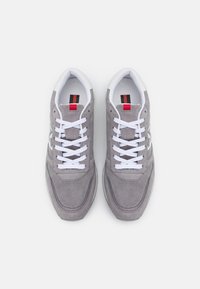 Gray suede sneakers with white laces, a padded tongue, and a logo detail. Features a white midsole and red accent on the tongue.