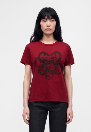 Jeune femme aux longs cheveux noirs porte un t-shirt rouge avec deux serpents noirs entrelacés et un texte délavé, associé à un pantalon noir.