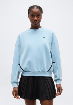 Femeie purtând un pulover albastru deschis cu margini negre și o fustă plisată neagră, stând în fața unui fundal alb simplu.