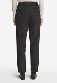 Svarta högmidjade byxor med texturerad tyg, elastiskt midjeband, två bakfickor och rak benmodell. Bärs med svarta skor.