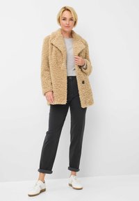 Beige nep-bont jas met een geknipt kraag, grote zwarte knopen, gedragen over een lichtgrijze top en zwarte manchetbroeken, gecombineerd met witte sneakers.