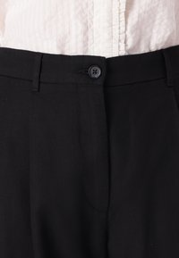 Schwarze Hose mit strukturiertem Finish, mit einem einzelnen Knopfverschluss, Gürtelschlaufen und seitlichen Taschen, die minimalistische Designdetails aufweisen.