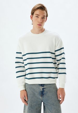 Weißes Sweatshirt mit dunkelblauen horizontalen Streifen. Mit Rundhalsausschnitt und gerippten Ärmelbündchen. Kombiniert mit hellblauen Jeans.