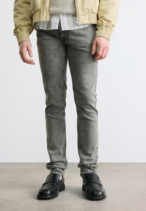 Bărbat purtând o jachetă bej deschis, pulover tricotat în straturi și cămașă cu dungi, blugi gri slim-fit și mocasini negri din piele, stând pe podea gri.