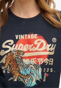 Mörkblå t-shirt med en broderad tiger och vågmotiv, med guld- och röd accenter, samt "SuperDry" text i krämfärg.