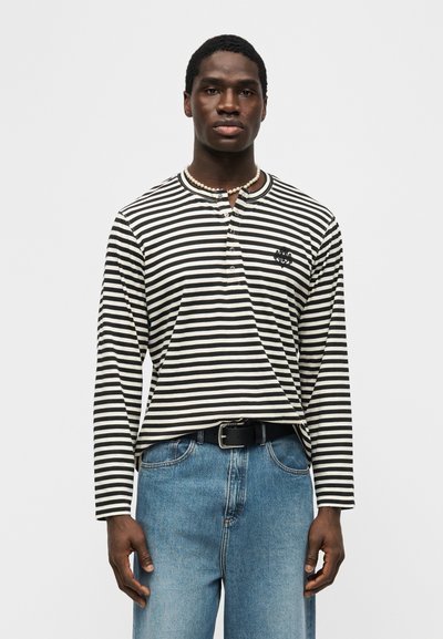 Άνδρας με μακρυμάνικο πουκάμισο ριγωτό σε ασπρόμαυρο χρώμα με μικρό λογότυπο στο στήθος, μπλε τζιν, μαύρη ζώνη και κολιέ με χάντρες, στέκεται μπροστά σε απλό φόντο.