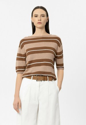 Frau mit glatten dunklen Haaren trägt einen braun-beigen gestreiften Pullover, weiße High-Waist-Hose und braunen Gürtel und steht vor weißem Hintergrund.