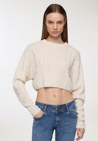 Szydełkowy, krótki sweter w kolorze off-white, z teksturowanym wzorem z kabli i diamentów. Połączony z wysokim stanem niebieskich dżinsów.