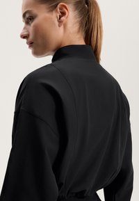 Černá košile s vysokým límcem, spadlými rameny a hladkou texturou. Design je minimalistický se střihem bez viditelných prvků.