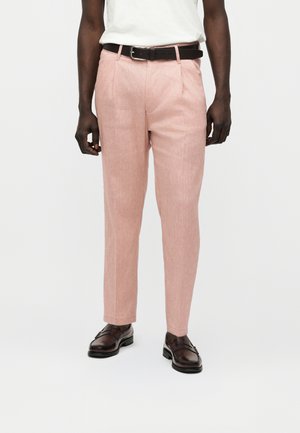 Mężczyzna w jasno-różowych spodniach, ciemnobrązowym pasku, białej koszuli i ciemnobrązowych eleganckich butach stojący na jednolitym tle.