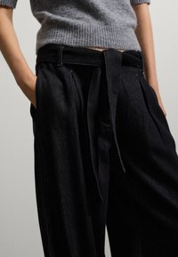 Persoon in hoge zwarte wijde broek met een stoffen riem en een ingestopt grijs brei trui, met handen in de zakken.