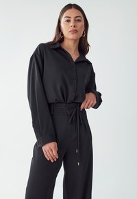 Black collared shirt with button placket, long sleeves, and relaxed fit, paired with high-waisted trousers featuring a fabric belt and tie detail.