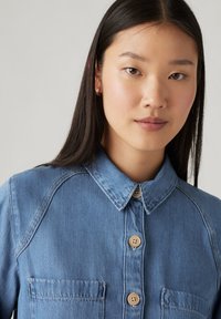 Light blue denim shirt featuring a classic collar, button-up front, two chest pockets, and subtle texture with curved seam details.