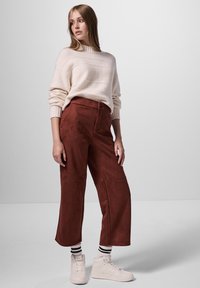 Beige tröja, hög krage, avslappnad passform. Rostfärgade mockabyxor, vida ben, framfickor. Vita sneakers med svarta och vita randiga strumpor.