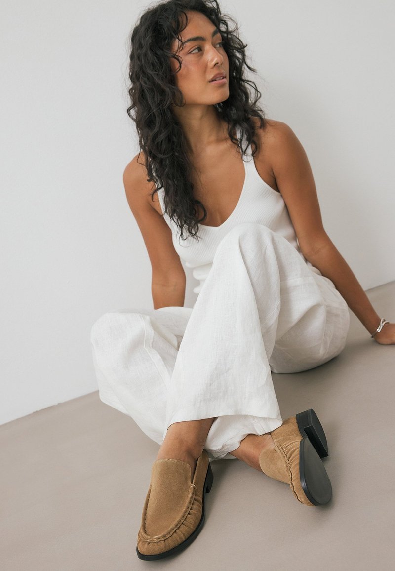 Braune Wildleder-Loafers mit gesticktem Akzent, ausgestattet mit einem niedrigen schwarzen Absatz, kombiniert mit weißen Leinen-Hose im weit geschnittenen Stil und einem weißen Tanktop.