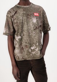 Mann trägt ein grün-braun gemustertes Diesel-T-Shirt mit rotem Logo-Patch auf der Brust, kombiniert mit dunkelbraunen Hosen, die Hände entspannt an den Seiten.