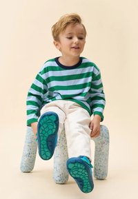 Camisa de rayas de manga larga en verde y azul claro, combinada con pantalones beige claros. Zapatos azules con suelas verdes texturizadas, sentados sobre un taburete abstracto.