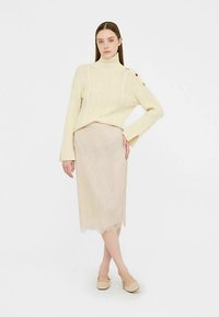 Jasnobeżowy dzianinowy golf z kablem i guzikami, zestawiony z jasną ołówkową spódnicą wykończoną koronką.