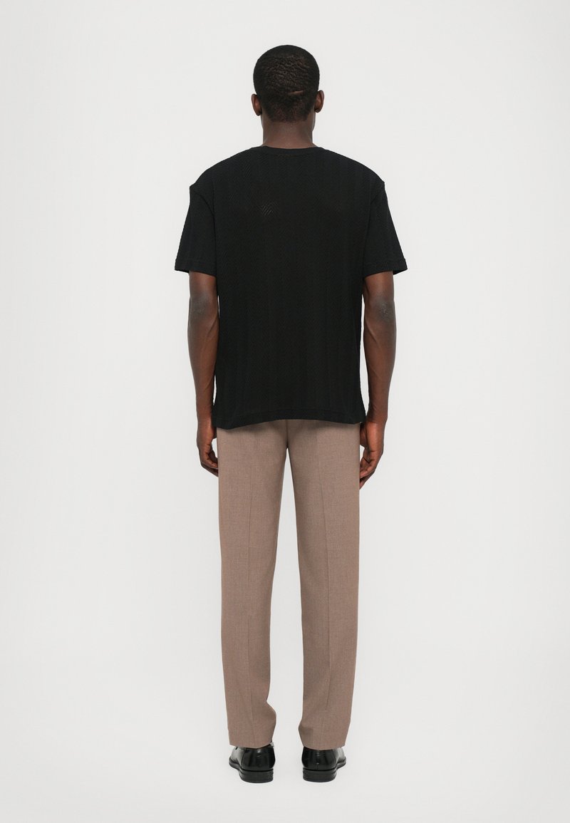 Black short-sleeve T-shirt with a textured pattern, paired with light brown trousers and black shoes, viewed from the back.