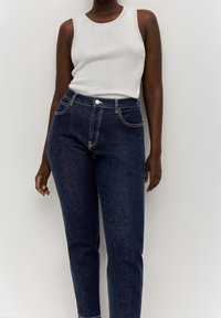 Femme portant un haut sans manches côtelé blanc et un jean en denim bleu foncé taille haute, debout contre un mur uni.