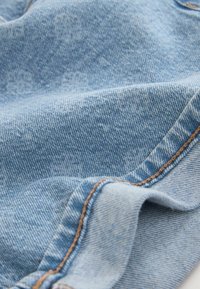 Tissu en denim bleu clair avec un tissage diagonal visible et des coutures marron le long d'une couture courbe.