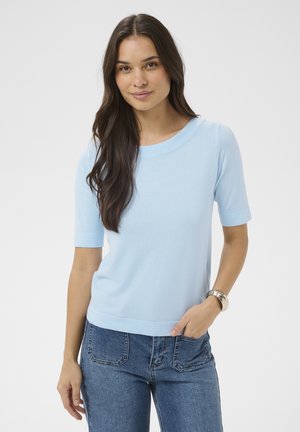 Donna con lunghi capelli scuri che indossa una maglietta azzurro chiaro a maniche corte e jeans, con una mano in tasca, in piedi contro uno sfondo bianco.