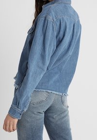 Kvinna som bär en ljusblå denimjacka med fransade kanter och matchande denimjeans, stående framför en enfärgad ljus bakgrund.