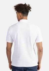 Valkoinen poolopaita, jossa on klassinen kaulus, lyhyet hihat ja sileä pinta, nähtynä takaa. Yksinkertainen muotoilu, ei näkyviä logoja tai kuvioita.