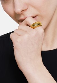 Anello d'oro, rotondo, con una superficie liscia e riflettente indossato sull'indice, accanto a una mano che copre la bocca. Abbigliamento nero visibile.