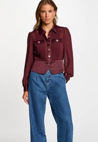 Blusa bordeaux con pieghe, dettagli a strisce e bottoni; abbinata a jeans blu larghi. Texture liscia e vestibilità su misura.