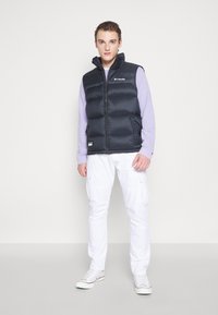 Černá péřová vesta s vysokým límcem, zapínání na zip, boční kapsy, nošena přes dlouhý fialový rukávový svetr, doplněna bílými cargo kalhotami a teniskami.