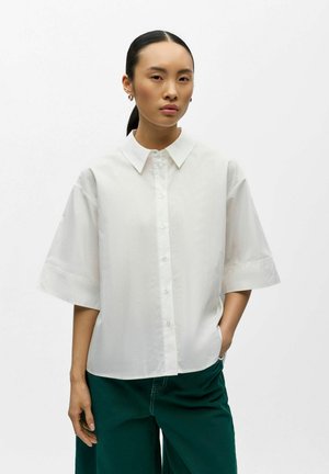 Kvinde med mørkt hår i lav hestehale iført en oversize hvid kortærmet skjorte med knapper og mørkegrønne bukser, står mod en ensfarvet baggrund.