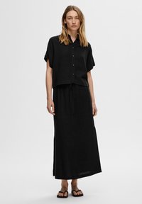 Chemise noire cropped à manches courtes, associée à une jupe longue noire avec une taille à cordon. Portée avec des sandales plates noires.