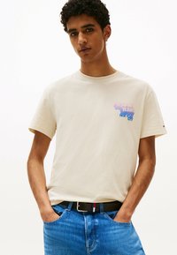 Beige bomulds T-shirt med korte ærmer og et flerfarvet "Tommy Jeans New York City" logo på brystet, parret med blå denim jeans.