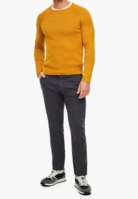 Senfgelber Strickpullover mit gerippter Textur, Rundhalsausschnitt, kombiniert mit dunkelgrauen Hosen und schwarz-weißen Sneakers. Einfaches Design, moderne Passform.