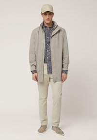 Chaqueta impermeable gris claro con un bolsillo con cremallera, llevada sobre una camiseta floral azul marino. Chinos beige y zapatillas a juego completan el conjunto.