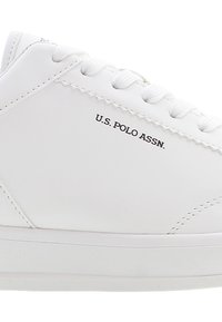 Bílé kožené tenisky s plochými bílými šňůrkami, šitými akcenty a černým logem "U.S. POLO ASSN." na boku. Hladký povrch s minimalistickým designem.