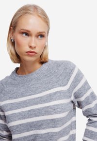 Maglione a righe grigie e bianche con colletto a giro e maniche raglan. Il modello ha i capelli biondi corti e indossa orecchini a forma di cuore in argento.