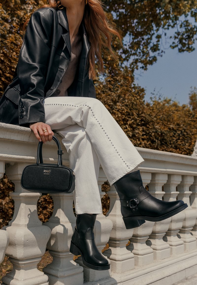 Woman wearing black leather jacket, white pants with studs, black ankle boots, and holding a black handbag while sitting on a stone railing outdoors.