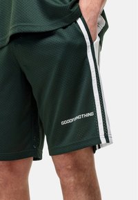 Zielone spodenki sportowe z przewiewnej siateczki, z białymi paskami po bokach i napisem "GOODFOR NOTHING" nadrukowanym na dolnej części przodu.