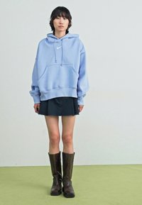 Sweatshirt azul claro com bolso frontal, capuz com cordão e logótipos brancos. Usado sobre uma saia mini preta e combinado com botas castanhas at�é ao joelho.