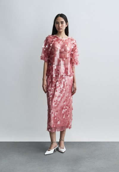 Robe pailletée rose à surface texturée, avec manches courtes et jupe longueur genou. Associée à des chaussures blanches à talons bas.