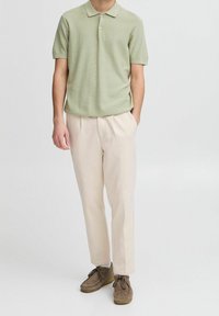 Man wearing light green short-sleeve polo, beige trousers, and brown suede lace-up shoes, standing with one hand in pocket on white background.