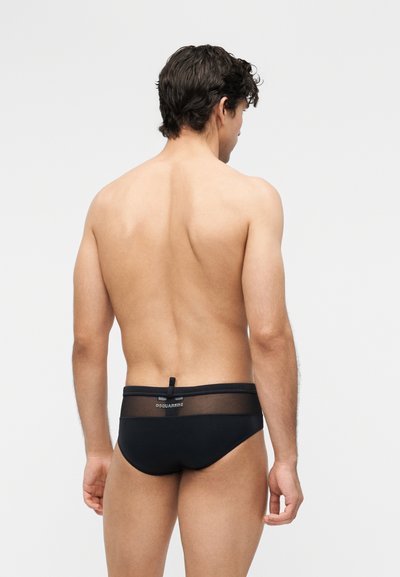 Άνδρας με σκούρα μαλλιά, φωτογραφημένος από πίσω, φορά μαύρα σλιπ με διάφανο ζωνάκι και το λογότυπο «DSQUARED2» σε απλό φόντο.